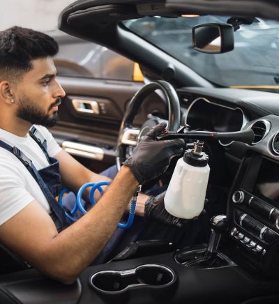 Detailer spraying interior cleaner inside a car while seated in the driver's seat